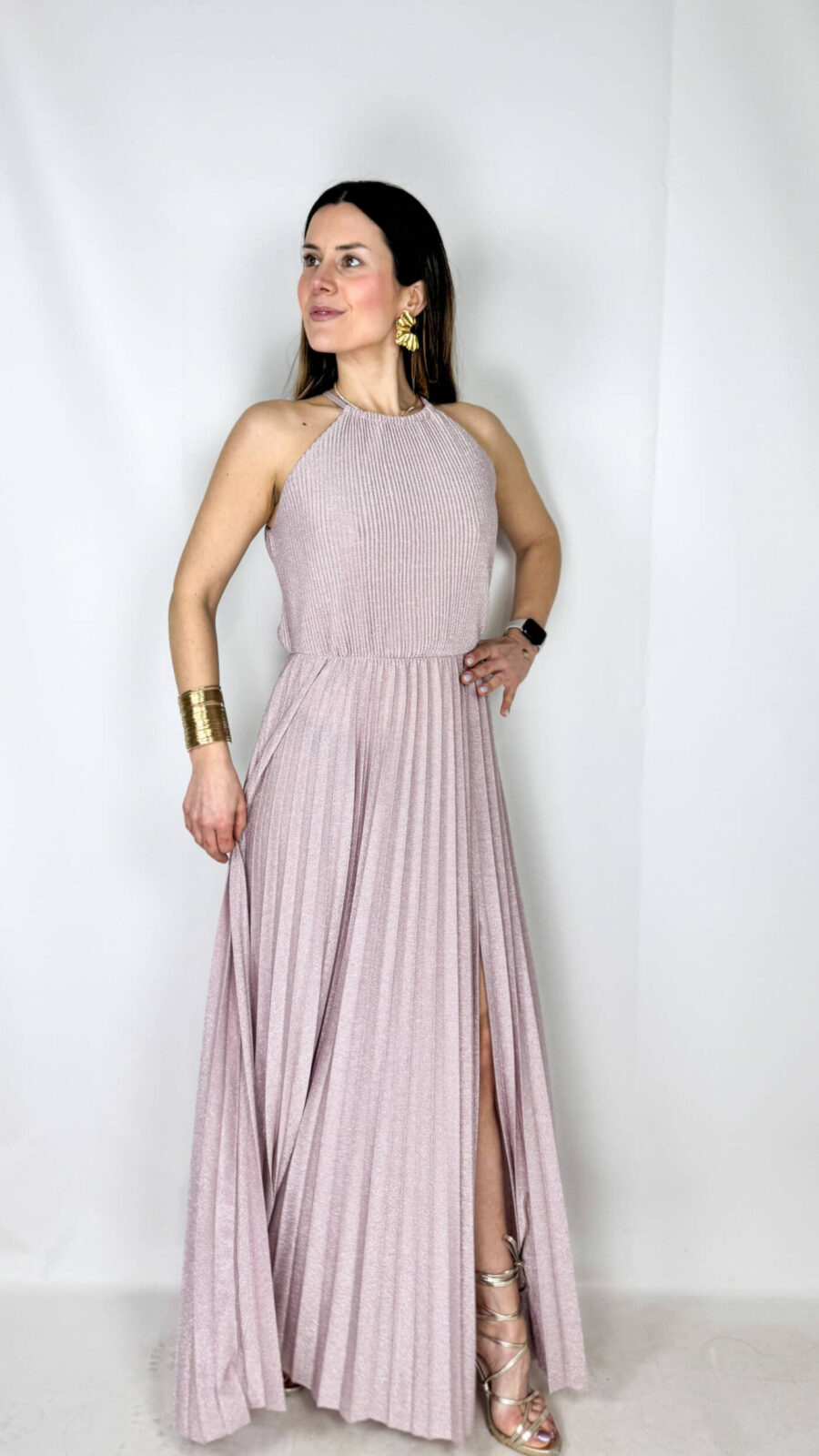 Abito da cerimonia lungo ed elegante, impreziosito da un raffinato tessuto brillantinato che illumina il look con discreta luminosità. Il modello presenta un girocollo alto con chiusura sul retro, che valorizza la linea del collo e dona un tocco sofisticato. Il corpetto plissettato crea movimento e leggerezza, mentre la gonna lunga fino alla caviglia è arricchita da un sensuale spacco che slancia la figura. L’elastico in vita garantisce comfort e una vestibilità adattabile, definendo delicatamente la silhouette. Perfetto per cerimonie, eventi eleganti e occasioni speciali.