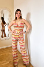 Tuta elasticizzata multicolore dallo stile vibrante e contemporaneo, ispirata alle iconiche fantasie zig-zag. Il design avvolgente valorizza la silhouette grazie al tessuto stretch che si adatta perfettamente al corpo, garantendo comfort e libertà di movimento. Il top a triangolo dona un’allure femminile e sensuale, mentre la schiena semi scoperta aggiunge un tocco audace e sofisticato, lasciando intravedere la pelle con eleganza. Il pantalone a gamba dritta completa il look con una linea fluida e slanciata, bilanciando perfettamente il fit aderente della parte superiore.