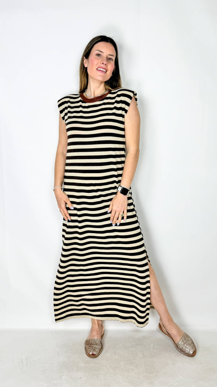 Vestito senza maniche dal design moderno e luminoso, caratterizzato da spalline leggermente imbottite che donano struttura e un tocco sofisticato alla silhouette. La fantasia a righe orizzontali aggiunge un effetto dinamico e contemporaneo, mentre il tessuto leggermente brillantinato regala eleganza e brillantezza discreta. Il modello girocollo e la vestibilità morbida assicurano comfort e leggerezza, rendendolo perfetto per diverse occasioni. Gli spacchi laterali completano il capo con un dettaglio femminile e arioso, ideale per valorizzare il movimento.