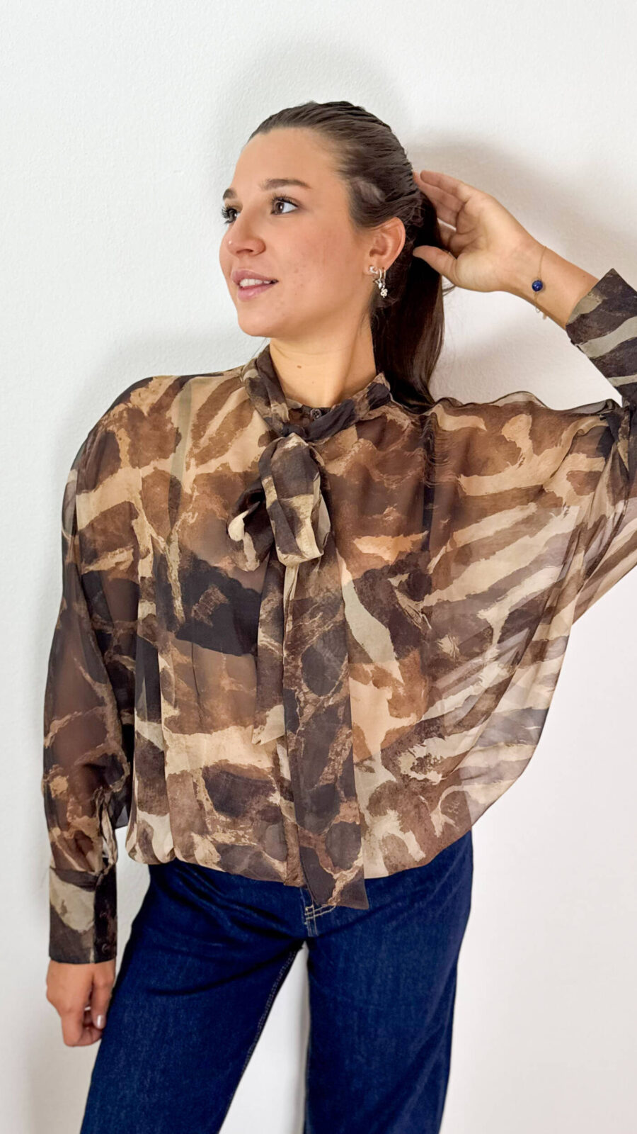 Delicata ma ricca di carattere, questa blusa in tessuto velato combina dettagli femminili e una linea contemporanea, creando un equilibrio perfetto tra eleganza e stile sportivo. La fantasia nei toni del marrone con effetto sfumato dona profondità al modello, mentre il fiocco sul collo aggiunge un dettaglio raffinato e distintivo. La vestibilità morbida accompagna la figura con naturalezza, senza segnare, rendendo la blusa piacevole da indossare e facile da abbinare. Le maniche ampie enfatizzano il volume del capo e sono completate da polsini con bottoni, per un tocco più curato e sofisticato. L'elastico sul fondo crea una linea leggermente raccolta, valorizzando la silhouette e conferendo alla blusa un aspetto moderno. Perfetta con pantaloni sartoriali o jeans, si adatta facilmente a diverse occasioni: elegante con un pantalone e un tacco, più casual e contemporanea con denim e sneakers.