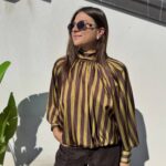 Eleva il tuo stile quotidiano o da ufficio con questa sofisticata blusa. Realizzata in un tessuto satinato di alta qualità che cattura splendidamente la luce, presenta un audace motivo a righe verticali nei toni del bordeaux e del bianco crema, creando un effetto allungante e moderno. I dettagli fanno la differenza: il collo alto drappeggiato conferisce un'aria bon-ton e sofisticata, mentre le voluminose maniche a palloncino, rifinite con polsini alti e abbottonati, aggiungono un tocco drammatico e femminile. La vestibilità è morbida sul busto e si raccoglie morbidamente sul fondo grazie a un elastico nascosto, creando un elegante effetto blusante che definisce il punto vita senza stringere