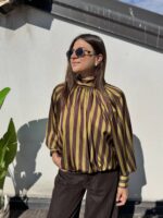 Eleva il tuo stile quotidiano o da ufficio con questa sofisticata blusa. Realizzata in un tessuto satinato di alta qualità che cattura splendidamente la luce, presenta un audace motivo a righe verticali nei toni del bordeaux e del bianco crema, creando un effetto allungante e moderno. I dettagli fanno la differenza: il collo alto drappeggiato conferisce un'aria bon-ton e sofisticata, mentre le voluminose maniche a palloncino, rifinite con polsini alti e abbottonati, aggiungono un tocco drammatico e femminile. La vestibilità è morbida sul busto e si raccoglie morbidamente sul fondo grazie a un elastico nascosto, creando un elegante effetto blusante che definisce il punto vita senza stringere