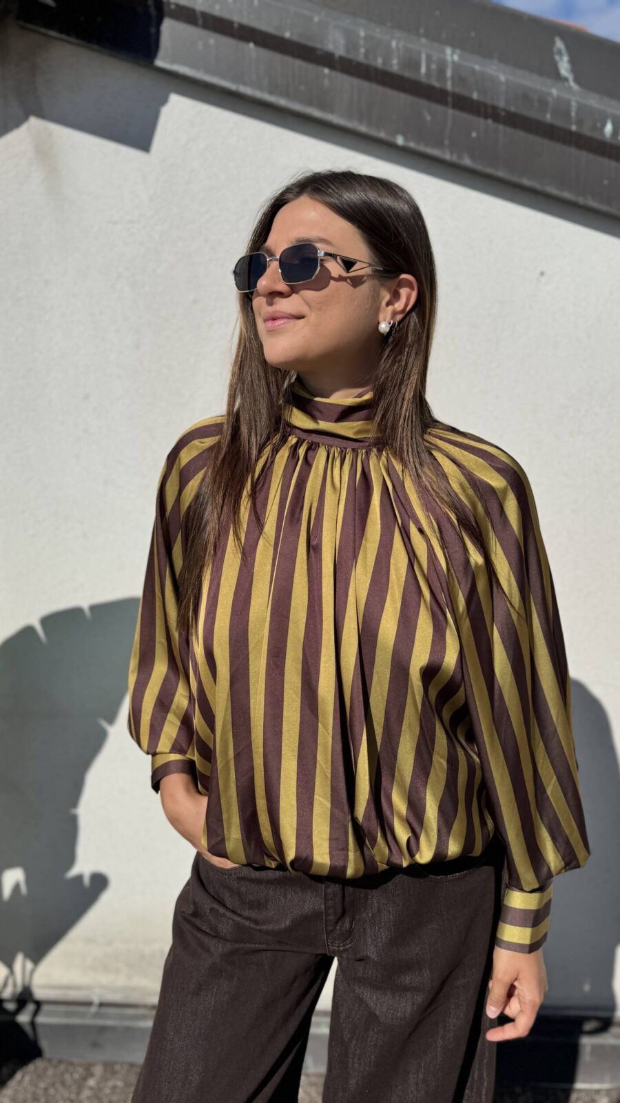 Eleva il tuo stile quotidiano o da ufficio con questa sofisticata blusa. Realizzata in un tessuto satinato di alta qualità che cattura splendidamente la luce, presenta un audace motivo a righe verticali nei toni del bordeaux e del bianco crema, creando un effetto allungante e moderno. I dettagli fanno la differenza: il collo alto drappeggiato conferisce un'aria bon-ton e sofisticata, mentre le voluminose maniche a palloncino, rifinite con polsini alti e abbottonati, aggiungono un tocco drammatico e femminile. La vestibilità è morbida sul busto e si raccoglie morbidamente sul fondo grazie a un elastico nascosto, creando un elegante effetto blusante che definisce il punto vita senza stringere