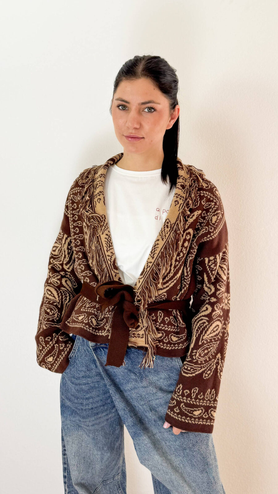 Aggiungi un tocco avvolgente e bohémien al tuo guardaroba con questo cardigan corto dalla finitura etnica. Caratterizzato da un elegante collo a revers impreziosito da dettagli sfrangiati, questo modello aperto sul davanti si distingue per la chiusura con coulisse in vita che permette di sagomare la silhouette a piacimento. Il ricco motivo jacquard su toni caldi del marrone e beige rende ogni outfit raffinato e contemporaneo, perfetto da abbinare a denim a vita alta o pantaloni sartoriali.