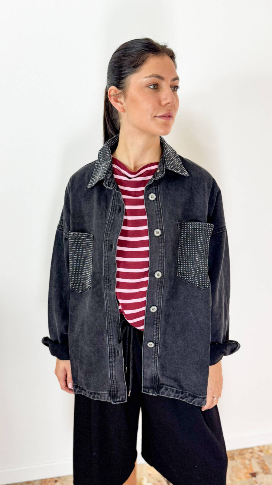 Descrizione Prodotto Eleva il tuo look street-style con questa versatile giacca camicia (overshirt) in denim nero dal lavaggio vintage. Caratterizzata da una vestibilità morbida ed oversize, è impreziosita da punti luce di strass che decorano le tasche e la trama del tessuto, regalandole un tocco glam ideale sia di giorno che di sera. Il taglio lungo stile camicia presenta il classico colletto, abbottonatura frontale con bottoni metallici e ampie tasche applicate sul petto. Perfetta da indossare sbottonata come capospalla leggero su t-shirt e top, oppure chiusa come statement piece sopra pantaloni a gamba larga o mini gonna. Dettagli Pr