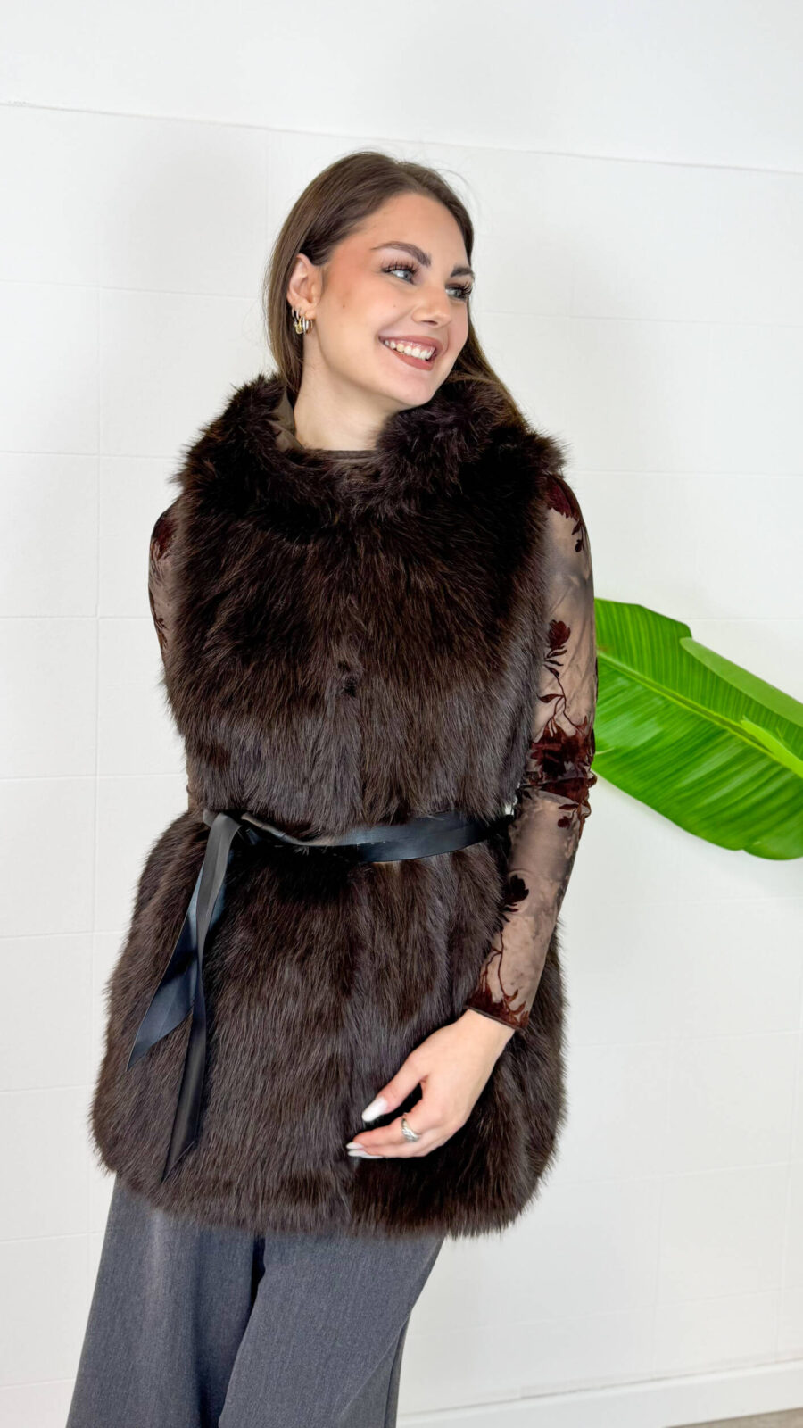 Aggiungi un tocco di raffinatezza e calore ai tuoi outfit invernali con questo gilet smanicato in ecopelliccia marrone scuro. Un capo morbido, avvolgente ed estremamente versatile, ideale per affrontare le giornate più fredde con stile. Dotato di un pratico cappuccio integrato e completamente foderato internamente, garantisce massimo comfort e protezione dal vento. Design e Silhouette: Taglio medio-lungo valorizzato da una cintura nera in vita che definisce la figura regalando una linea elegante e femminile. Dettagli Funzionali: Ampio cappuccio foderato e finitura in eco-pelliccia sintetica ad alta densità, morbida al tatto e dal look lussuoso. Come Abbinarlo: Perfetto indossato sopra una maglia a maniche lunghe, un dolcevita o una camicia trasparente per le serate speciali, abbinato a pantaloni a gamba larga o gonne midi.